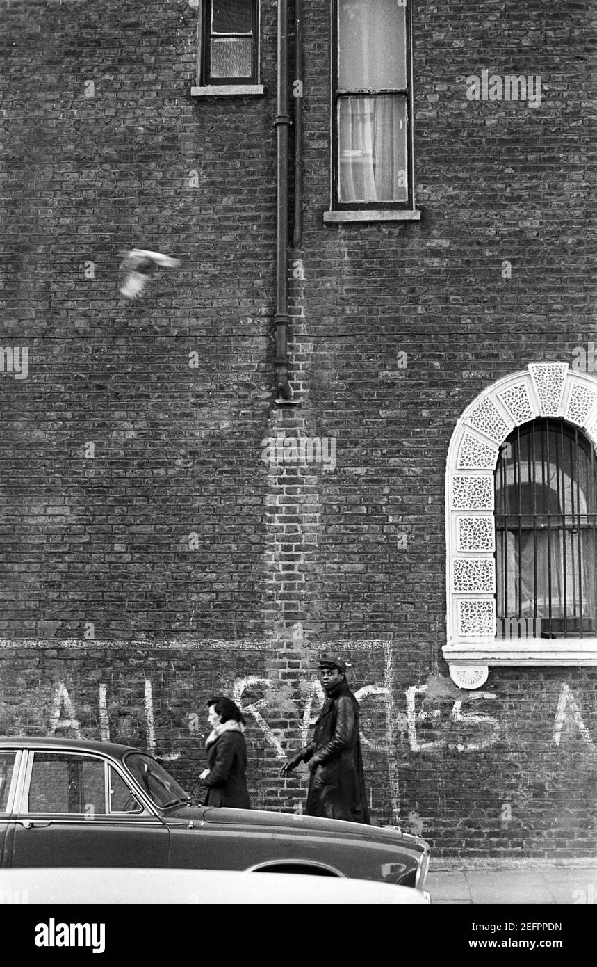 UK, West London, Notting Hill, 1973. Una donna bianca e un uomo nero camminano oltre i graffiti che recita 'tutte le razze....' scritto sulle pareti di Colville Houses, cul-de-sac, ora demolito e ricostruito. Foto Stock