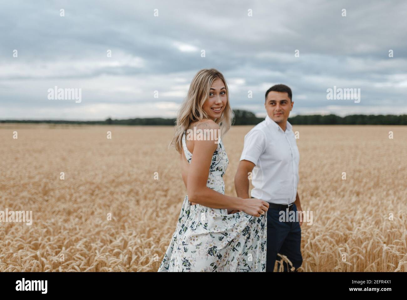 La donna sorridente e l'uomo attraversano un campo di grano che tiene le mani. Amanti del campo di grano Foto Stock