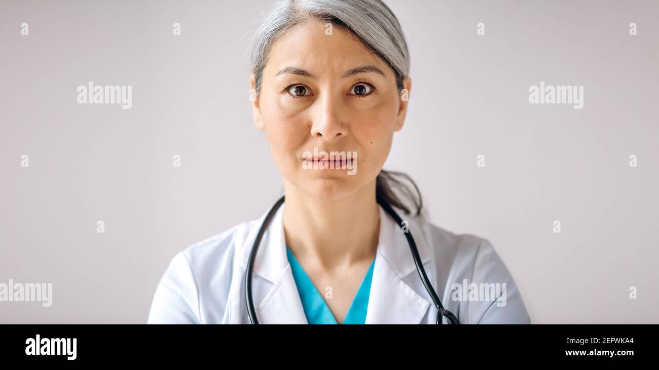 Ritratto panoramico di una dottoressa, medico o pediatra asiatica con capelli grigi in uniforme medica e stetoscopio, in piedi su uno sfondo isolato e guardando direttamente la fotocamera Foto Stock