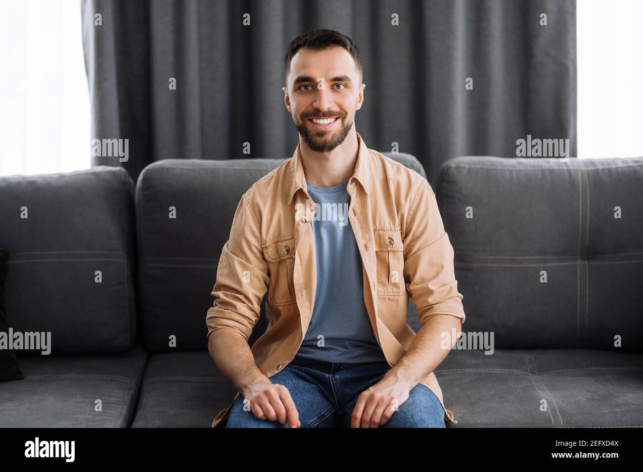 Ritratto di un attraente ragazzo caucasico vestito in elegante abbigliamento casual. Uomo sicuro guarda direttamente la macchina fotografica con un sorriso amichevole mentre si siede sul divano in un soggiorno Foto Stock