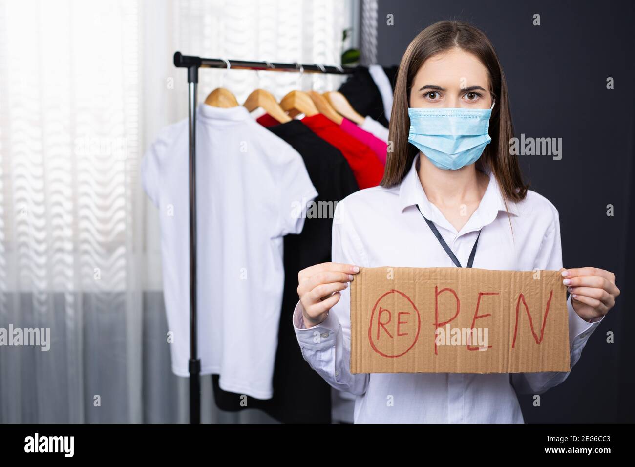 Ritratto di un bel proprietario di un negozio di abbigliamento con un cartello, cartello riaperto, aperto. Il venditore indossa una maschera protettiva nella pandemia del coronavirus Foto Stock
