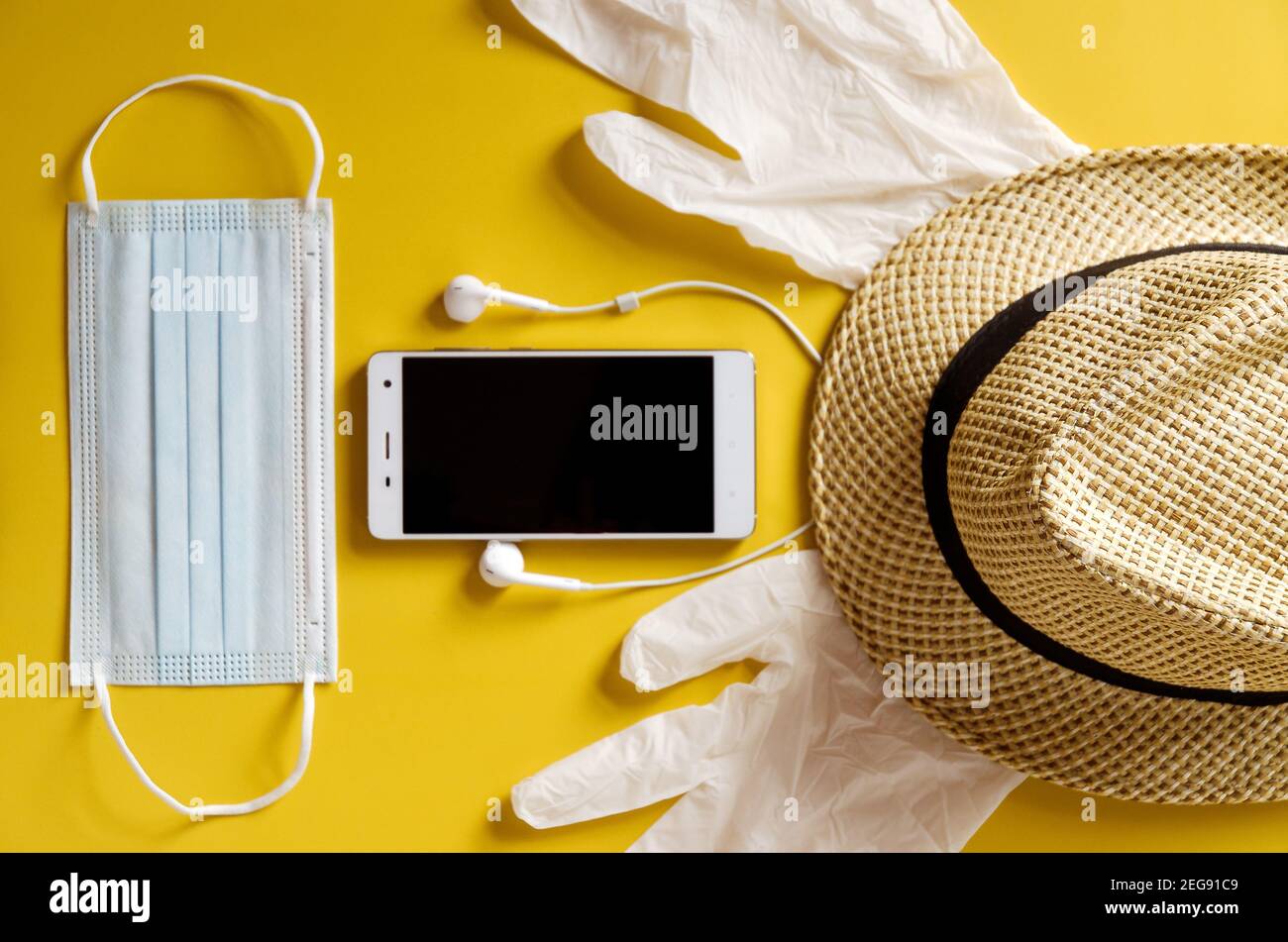 Viaggio dopo Covid-19 e nuovi concetti normali. Vista dall'alto della maschera medica, guanti, cuffie con telefono e cappello da spiaggia su sfondo giallo vicino u Foto Stock