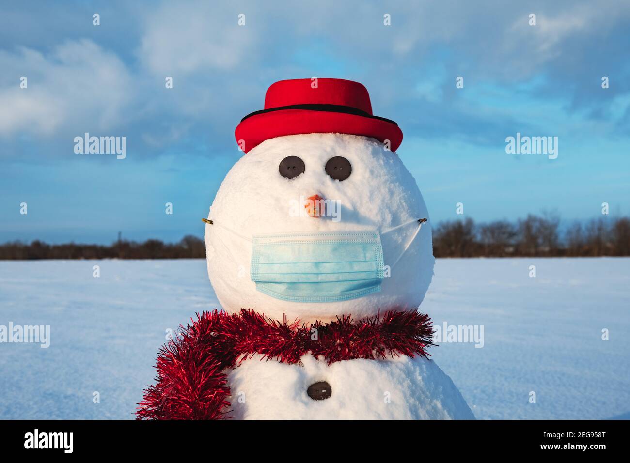 Simpatico pupazzo di neve in elegante cappello rosso con maschera medica su campo innevato. Concetto di allontanamento sociale e di viaggio annullato Foto Stock
