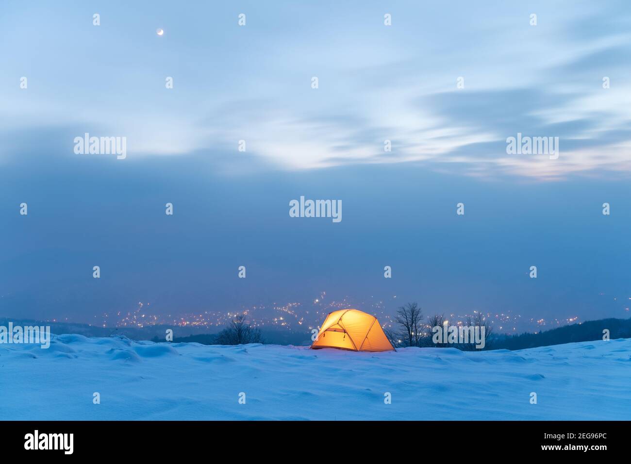 Tenda gialla illuminata dall'interno sullo sfondo di luci della città incandescenti nella nebbia. Incredibile paesaggio innevato. Campo turistico in montagna d'inverno. Concetto di viaggio Foto Stock