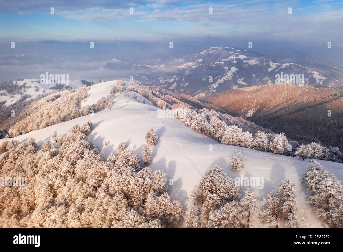 Incredibile vista aerea della catena montuosa, dei prati e delle cime innevate in inverno. Foresta con brina incandescente con luce calda luminosa alba Foto Stock