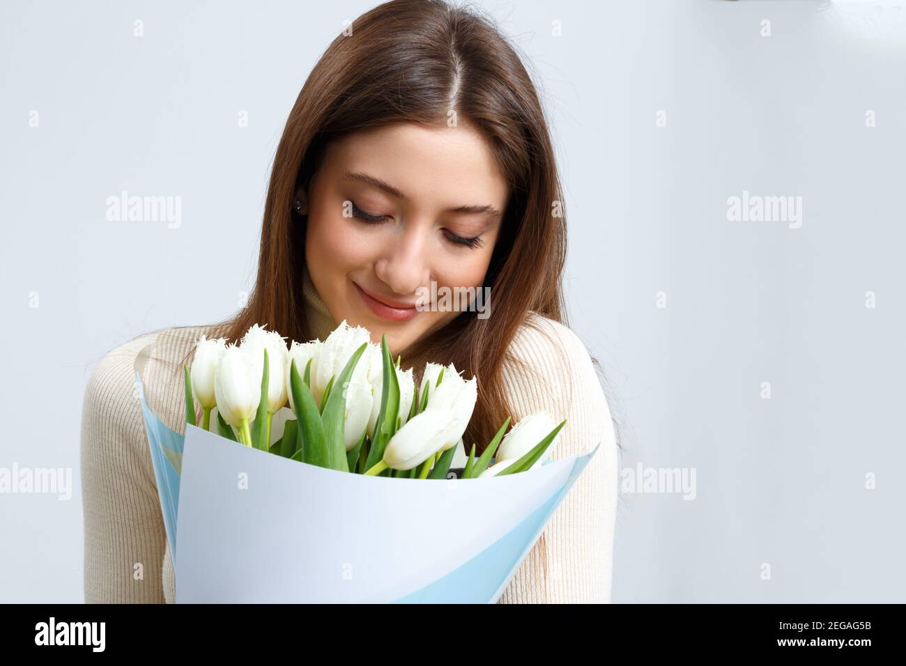 Closeup ritratto di giovane donna che tiene grande bouquet di tulipani bianchi su sfondo grigio. Una bella ragazza dai capelli castani chiuse gli occhi e mi respira Foto Stock