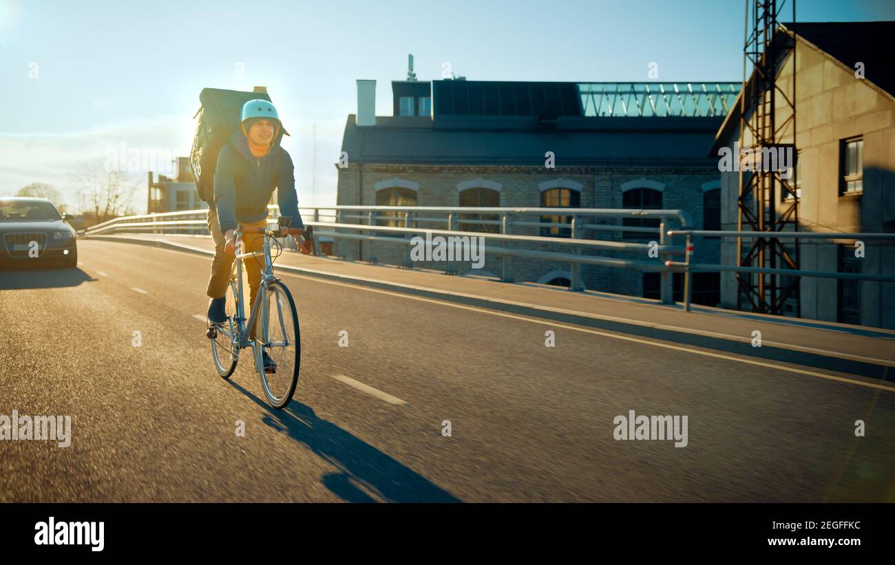 Happy Food Delivery Courier indossa lo zaino termico corre una bicicletta su strada per consegnare gli ordini per clienti e clienti. Giornata di sole nella città moderna con Foto Stock