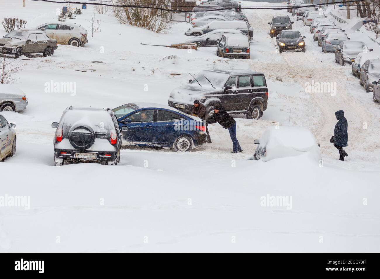 Tula, Russia - 13 febbraio 2021: Due uomini che spingono l'auto Hyundai Solaris bloccata attraverso un cortile innevato tra file di auto parcheggiate nella neve profonda in pendenza. Foto Stock