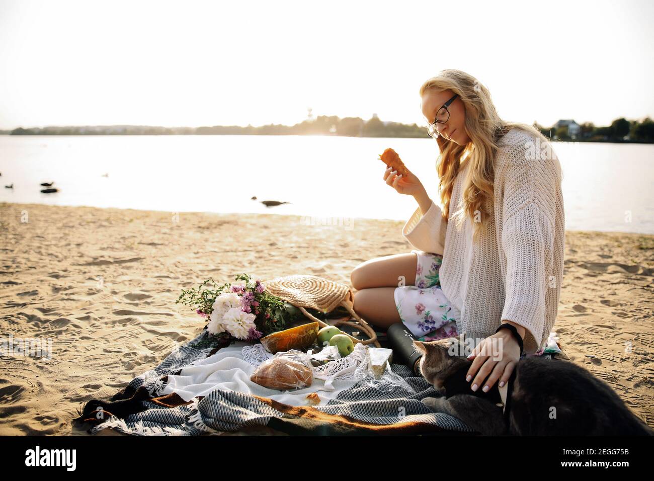 Giovane donna bionda sdraiata sulla spiaggia in autunno a. un pullover bianco lavorato a maglia con un gatto su un sole giornata di un picnic Foto Stock