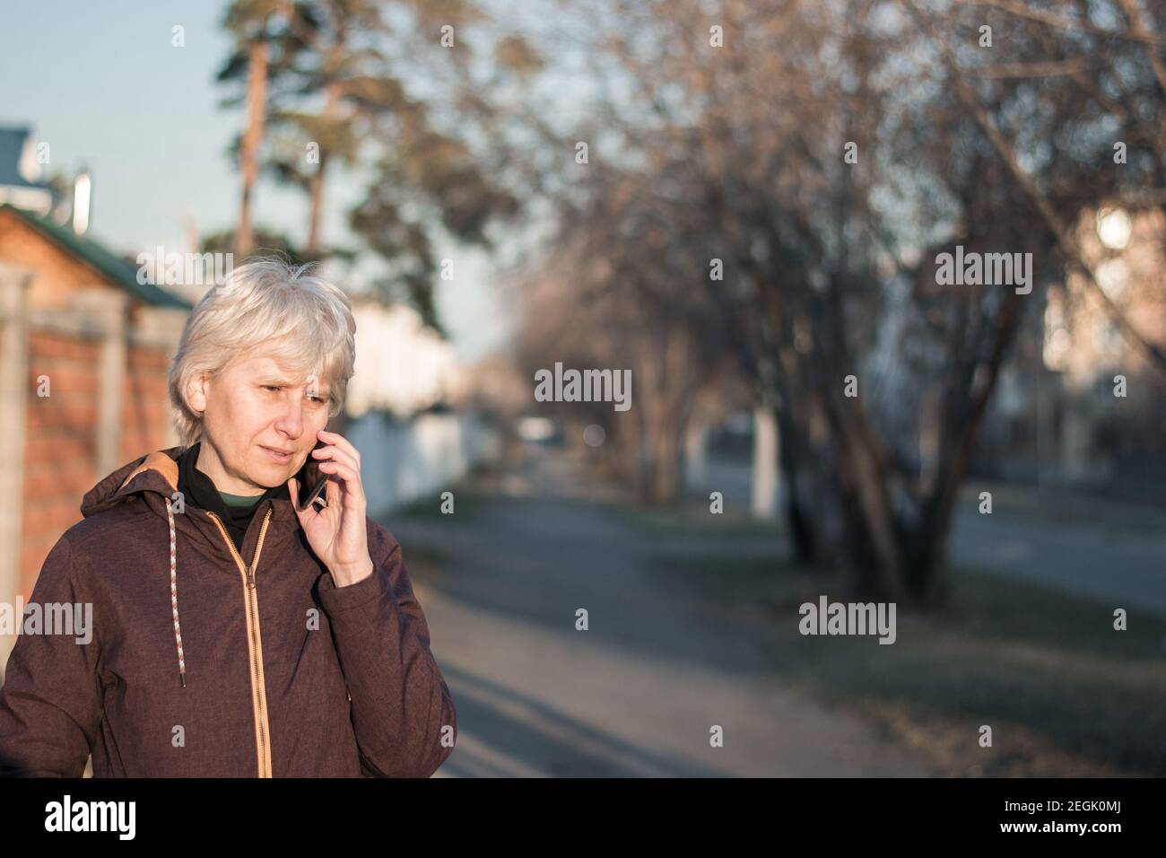 Una bella donna di mezza età in abiti casual sta parlando al telefono con un sorriso sul viso. Passeggiate per la città la sera al sole. Foto Stock