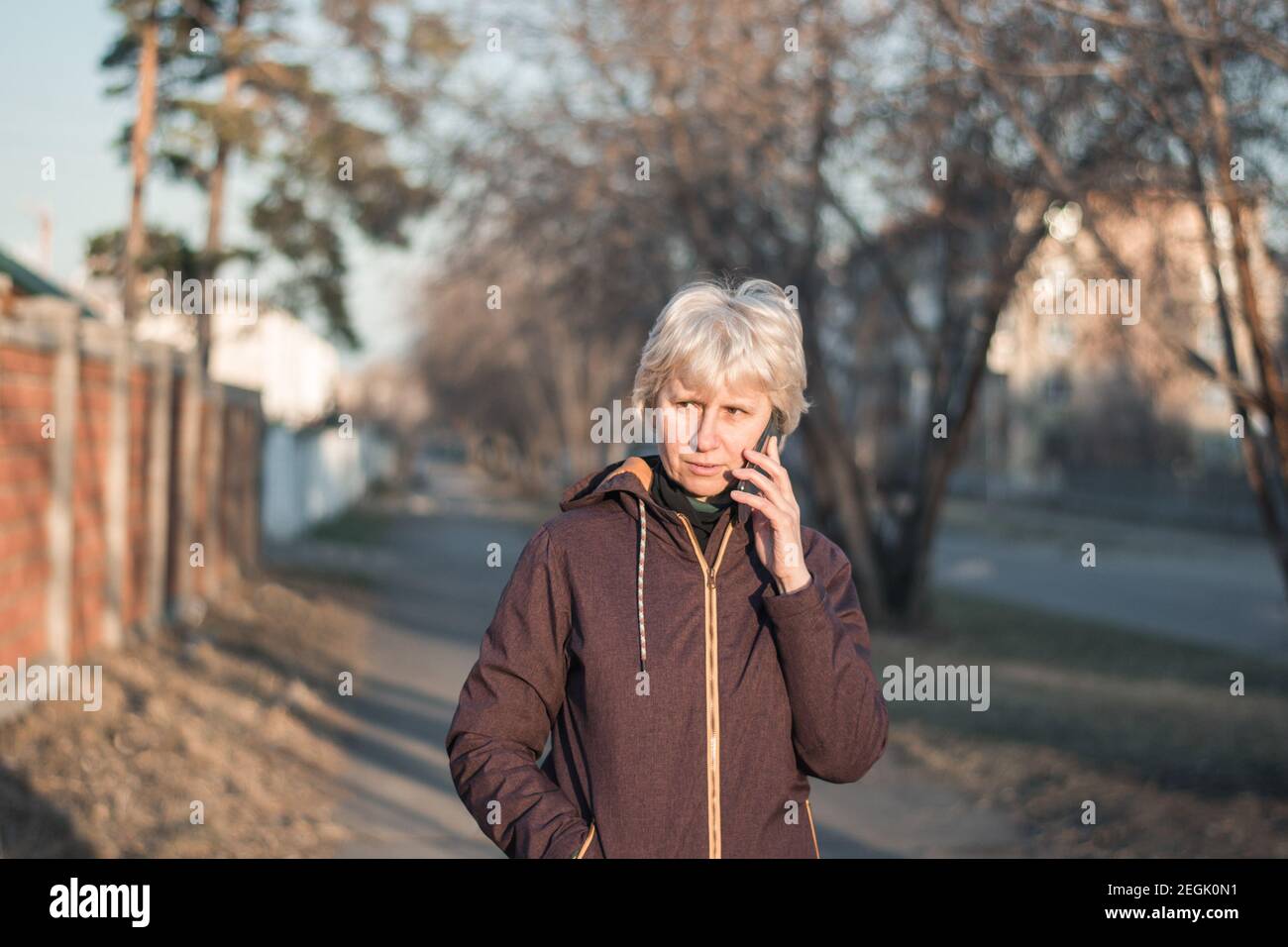 Una bella donna di mezza età in abiti casual sta parlando al telefono con un sorriso sul viso. Passeggiate per la città la sera al sole. Foto Stock
