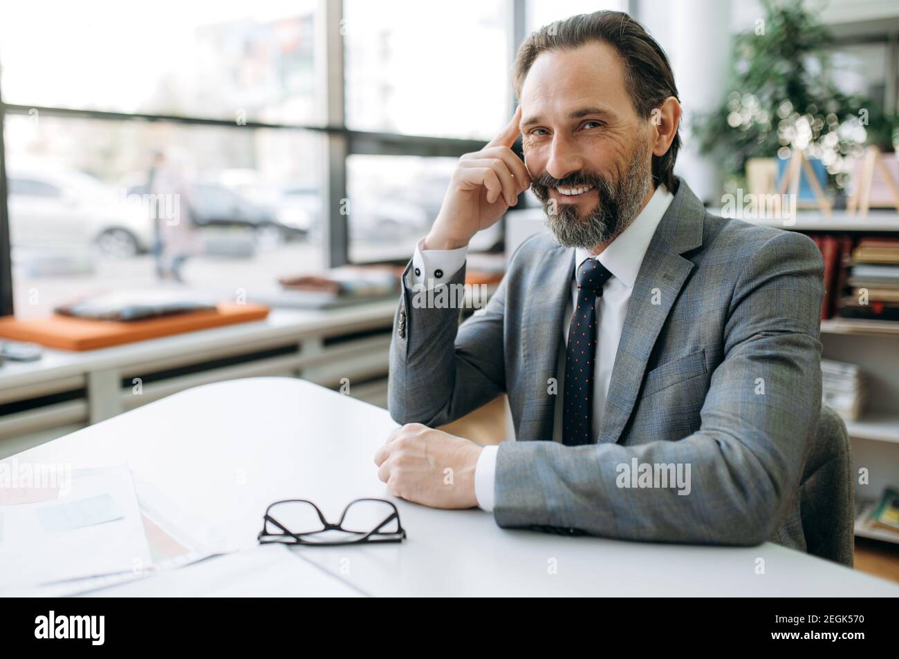 Ritratto di felice imprenditore di successo sul posto di lavoro. Un uomo d'affari sorridente di mezza età si siede alla scrivania, guarda la macchina fotografica, indossando un abito elegante e formale Foto Stock