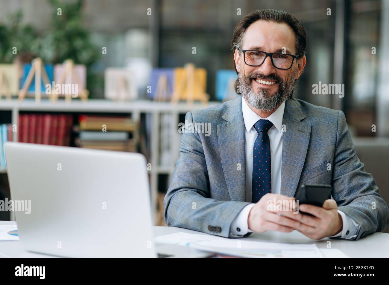 Un uomo d'affari sorridente dai capelli grigi guarda direttamente la telecamera, si siede alla scrivania. Felice dipendente maschio influente sta usando smartphone, e sorrisi amichevoli Foto Stock