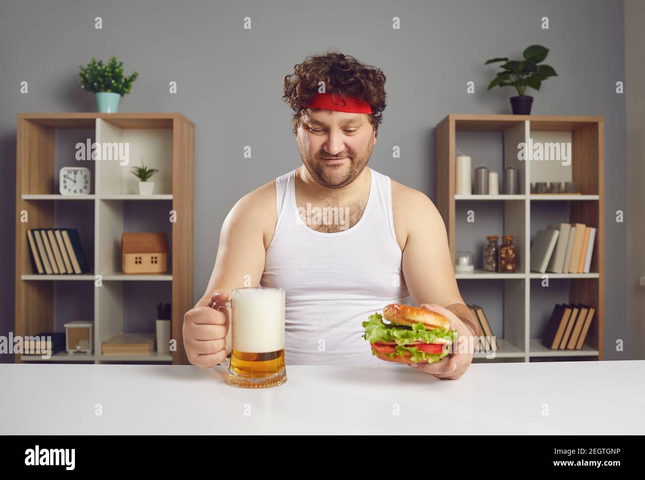 Felice giovane uomo seduto al tavolo, mangiando deliziosi cheeseburger e bevendo birra Foto Stock