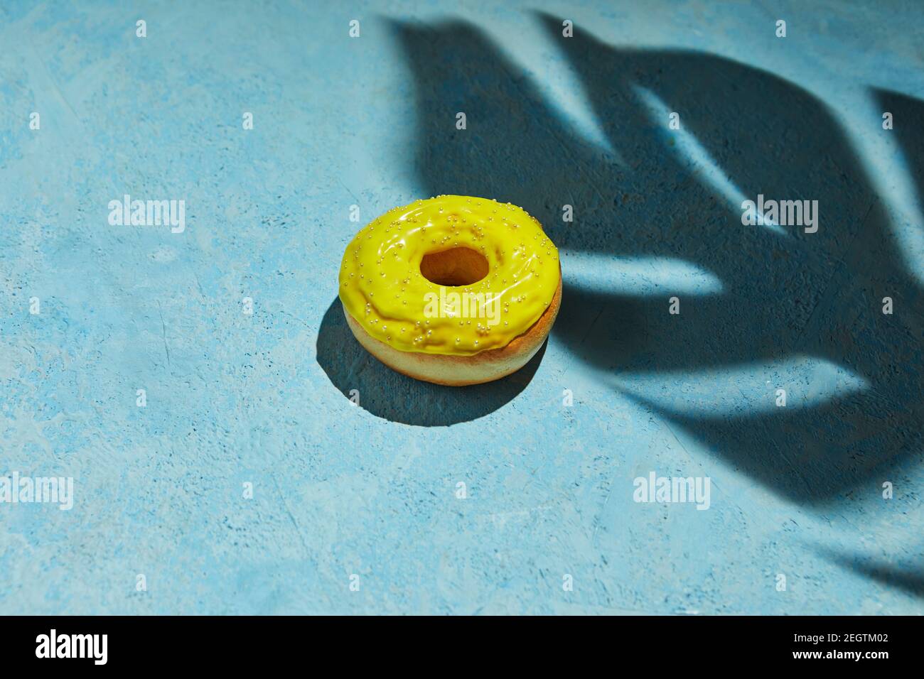 Ciambella con glassa gialla e spruzzi con ombra da foglia di monstera su sfondo blu. Foto Stock