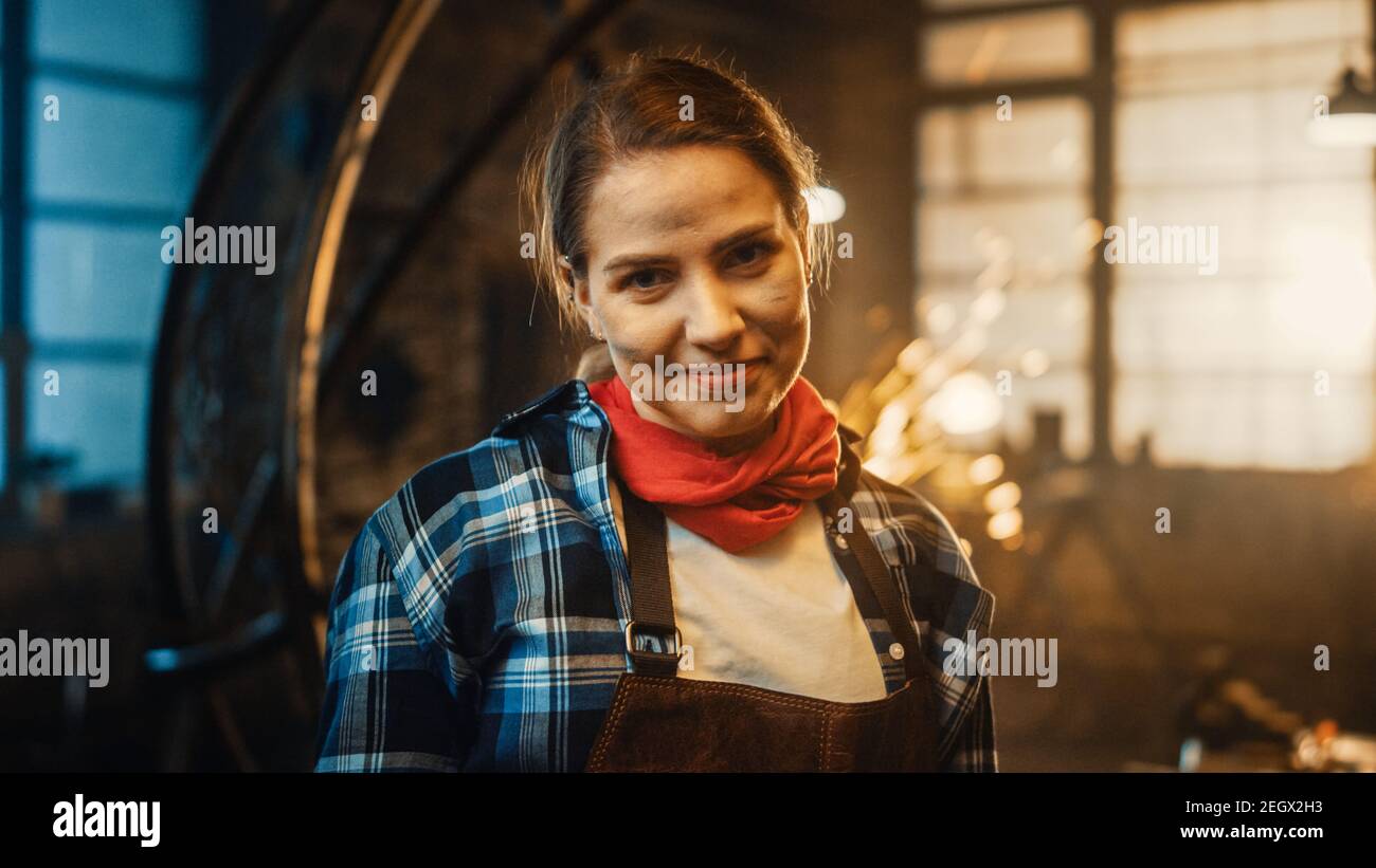 Giovane bella Empowerering Donna con l'orecchio Piercing sorride delicatamente alla macchina fotografica. Autentico Fabricator che indossa abiti da lavoro in un laboratorio di metallo. Scintille Foto Stock