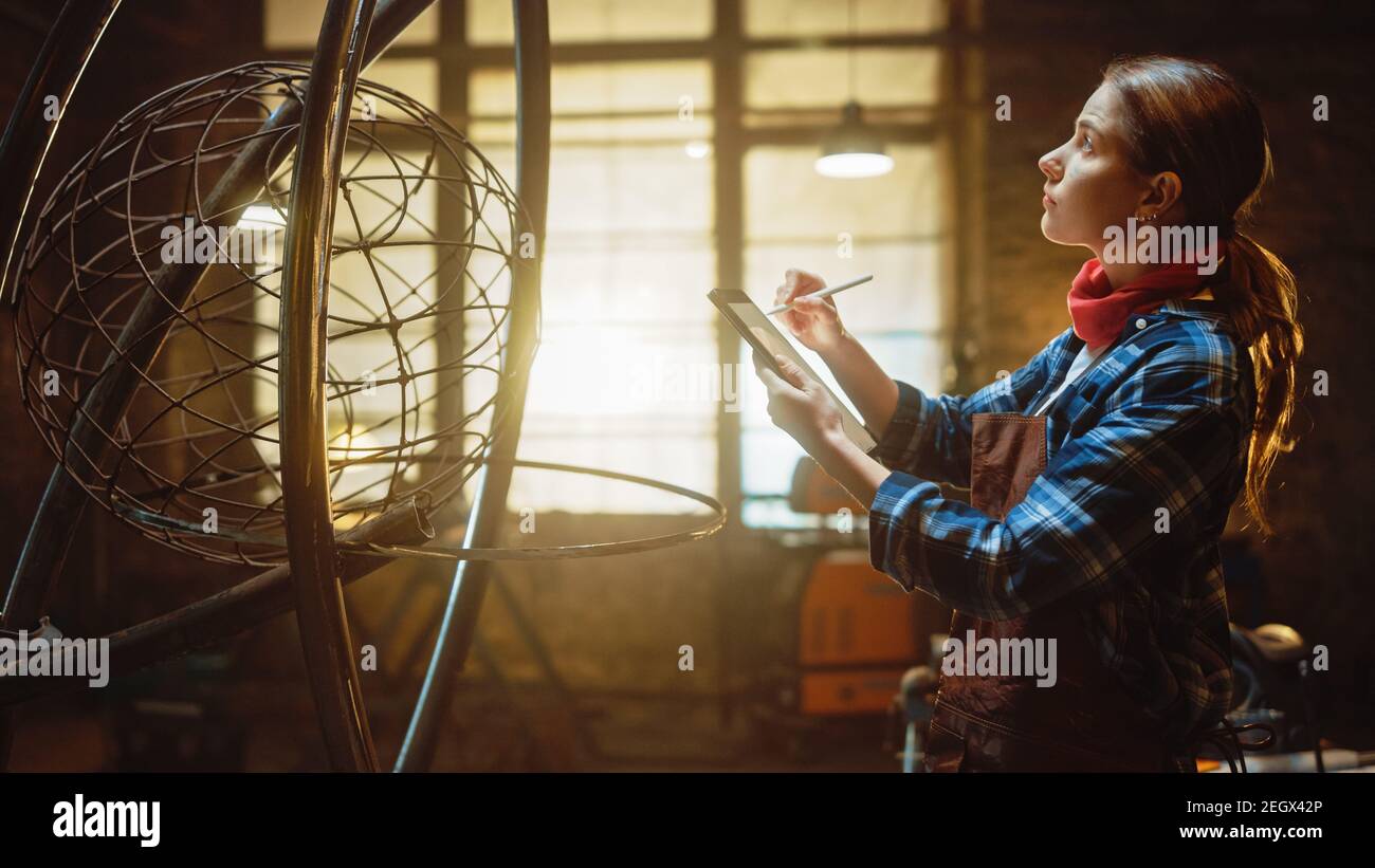 Bella artista femminile schizzi su un Tablet computer accanto a brutale scultura in metallo in Studio. Tomboy Girl indossa una camicia a scacchi e un grembiule Foto Stock