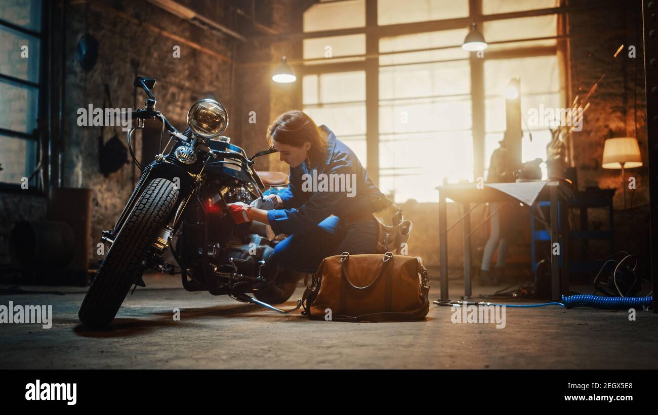 Giovane bella meccanica femminile sta lavorando su una Bobber personalizzata motocicletta. Ragazza di talento che indossa una tuta blu. Utilizza una chiave a cricchetto per stringere Foto Stock