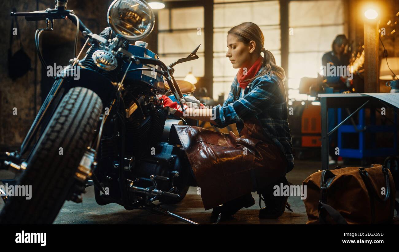 Giovane bella meccanica femminile sta lavorando su una Bobber personalizzata motocicletta. Ragazza di talento che indossa una camicia a scacchi e un grembiule. Utilizza uno Spanner per Foto Stock