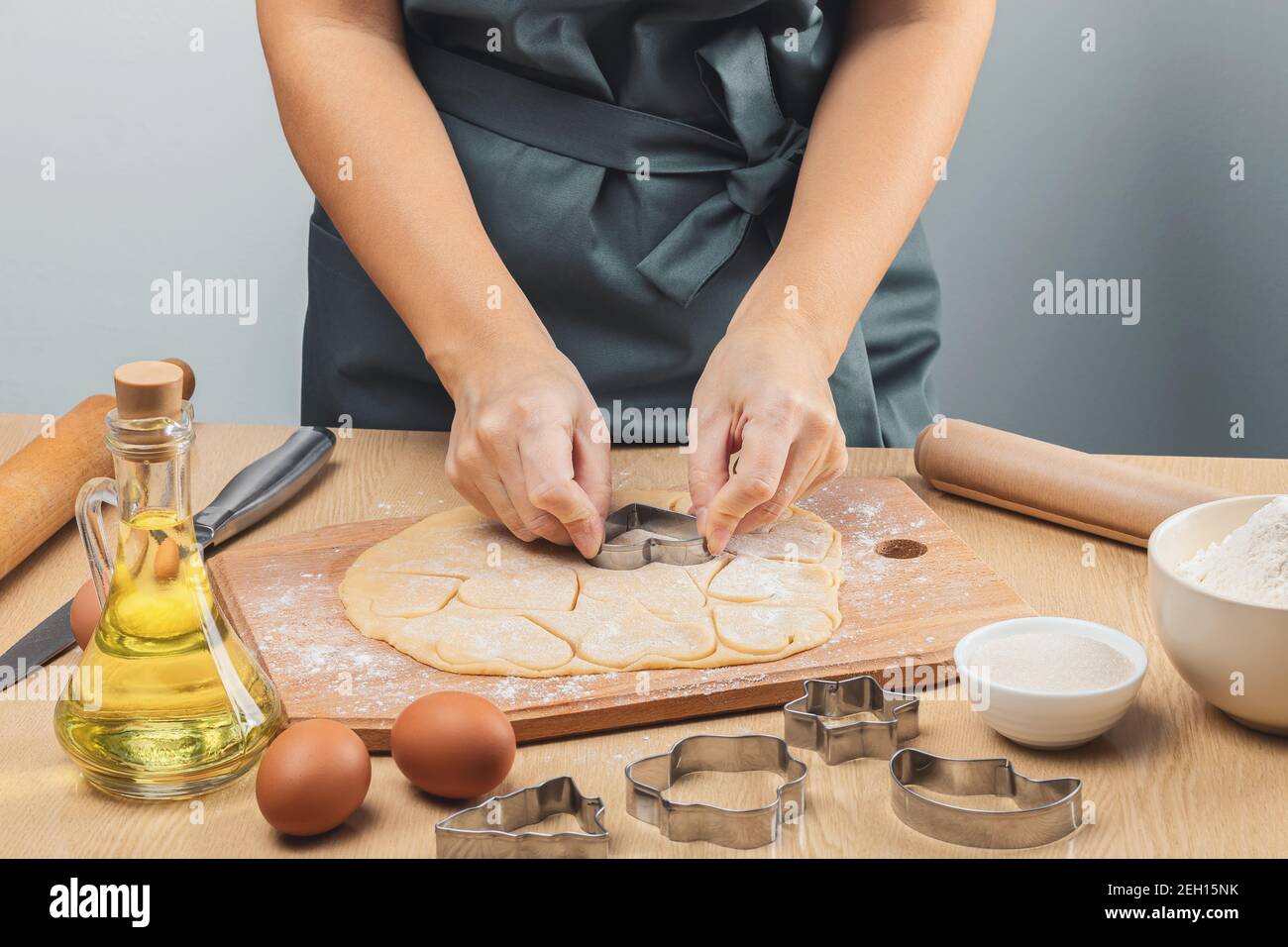 Una ragazza in un grembiule grigio senza viso taglia i cuori di pasta frolla con una muffa di ferro. Sul tavolo si trovano un laminino di legno, farina, olio in un gl Foto Stock