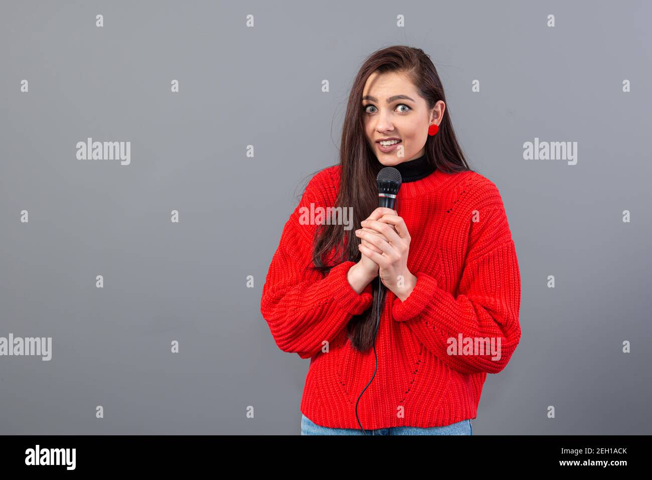giovane, emozionale brunetta con microfono vestito da un maglione rosso  canta il karaoke o dice un discorso, isolato su grigio Foto stock - Alamy