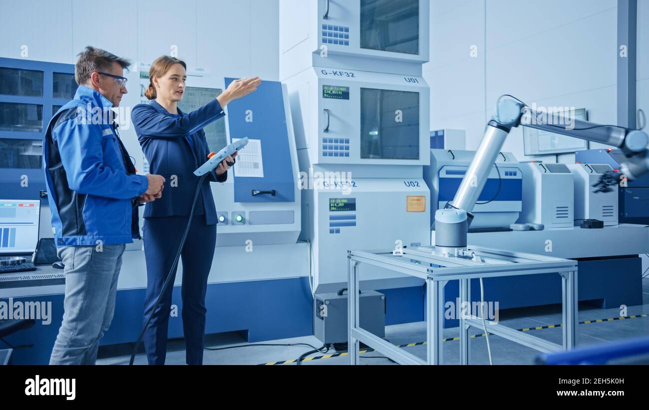 Fabbrica moderna: Responsabile di progetto femminile e ingegnere maschile in piedi, parlando e utilizzando Tablet computer per la programmazione della linea di assemblaggio del braccio robot Foto Stock