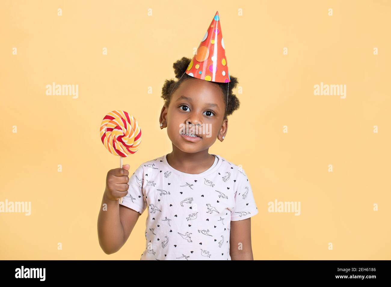 Primo piano di piccola adorabile afroamericana ragazza in cappello da festa che tiene lollipop dolce in mani, in posa per la fotocamera su sfondo giallo, Copy Space Foto Stock