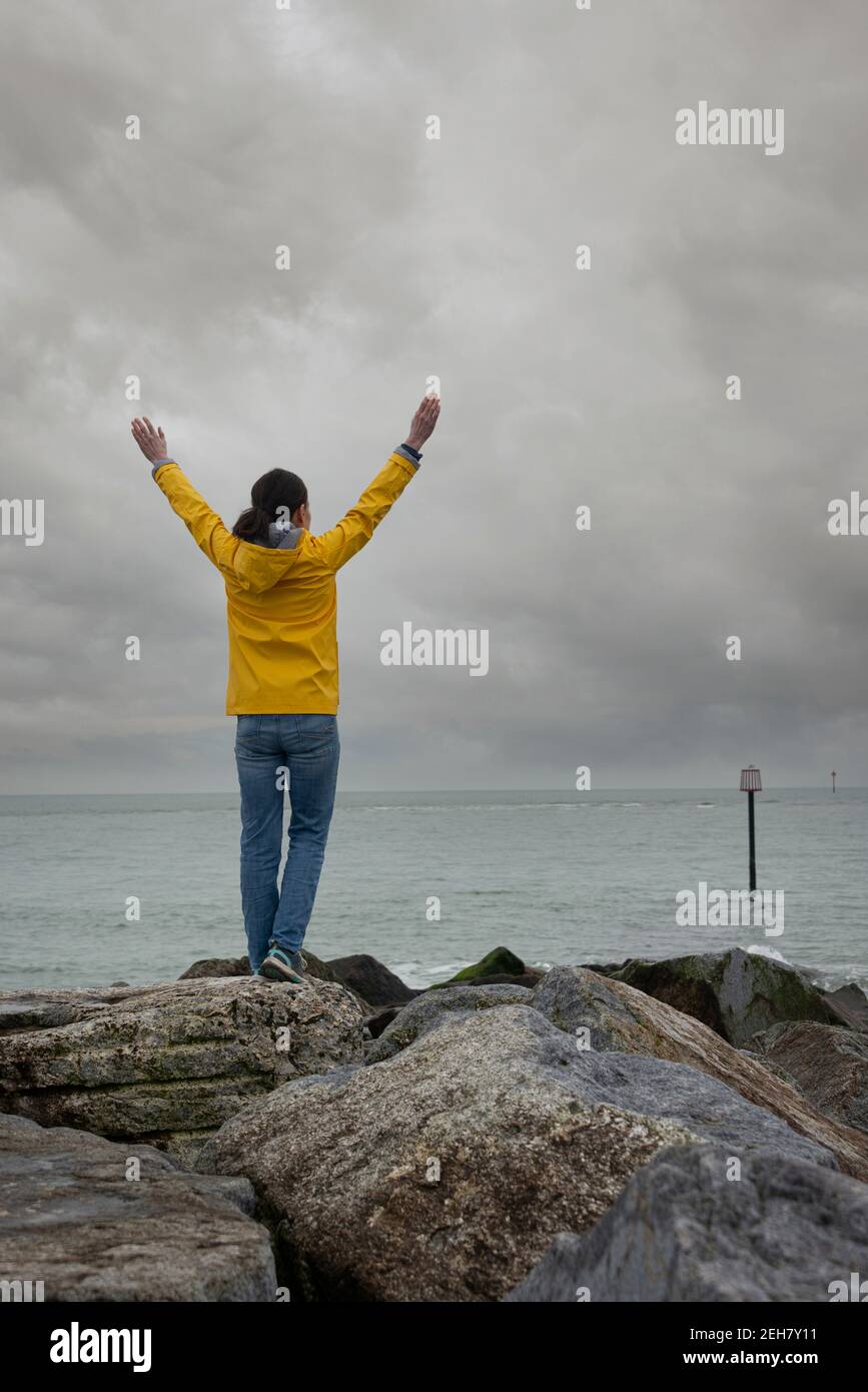 Vista posteriore di una donna in piedi su rocce con le braccia sollevate guardando verso il mare in una giornata grigia. Foto Stock