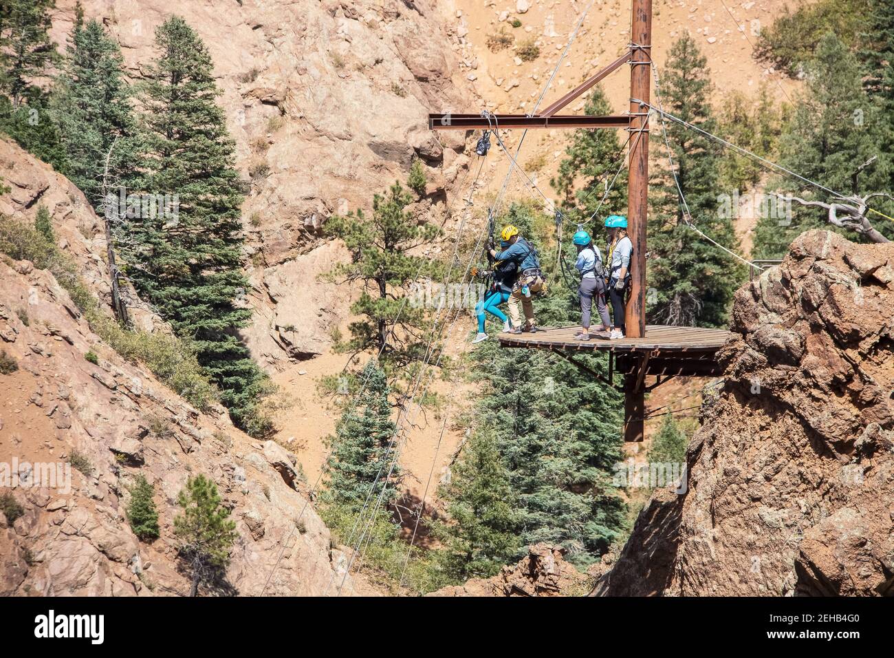 Persone su una piattaforma in montagna che si preparano a. rappel giù - uomo che aiuta la donna in imbracatura come lei si prepara a partire mentre gli altri guardano Foto Stock