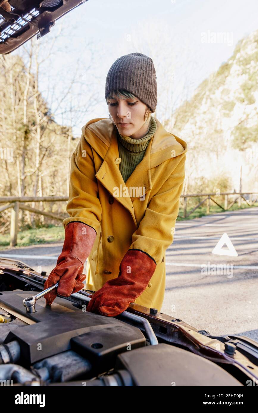 Giovane donna in abiti caldi e guanti non conduttivi morsetti sulla batteria dell'auto sotto il cofano nel soleggiato parcheggio autunnale Foto Stock