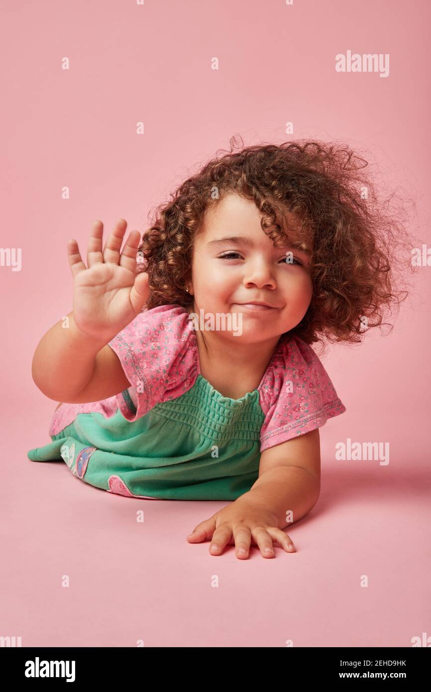 Adorabile bambino in abito con capelli ricci che guarda la fotocamera si appoggia con le mani sul pavimento dicendo addio con la mano gesto Foto Stock