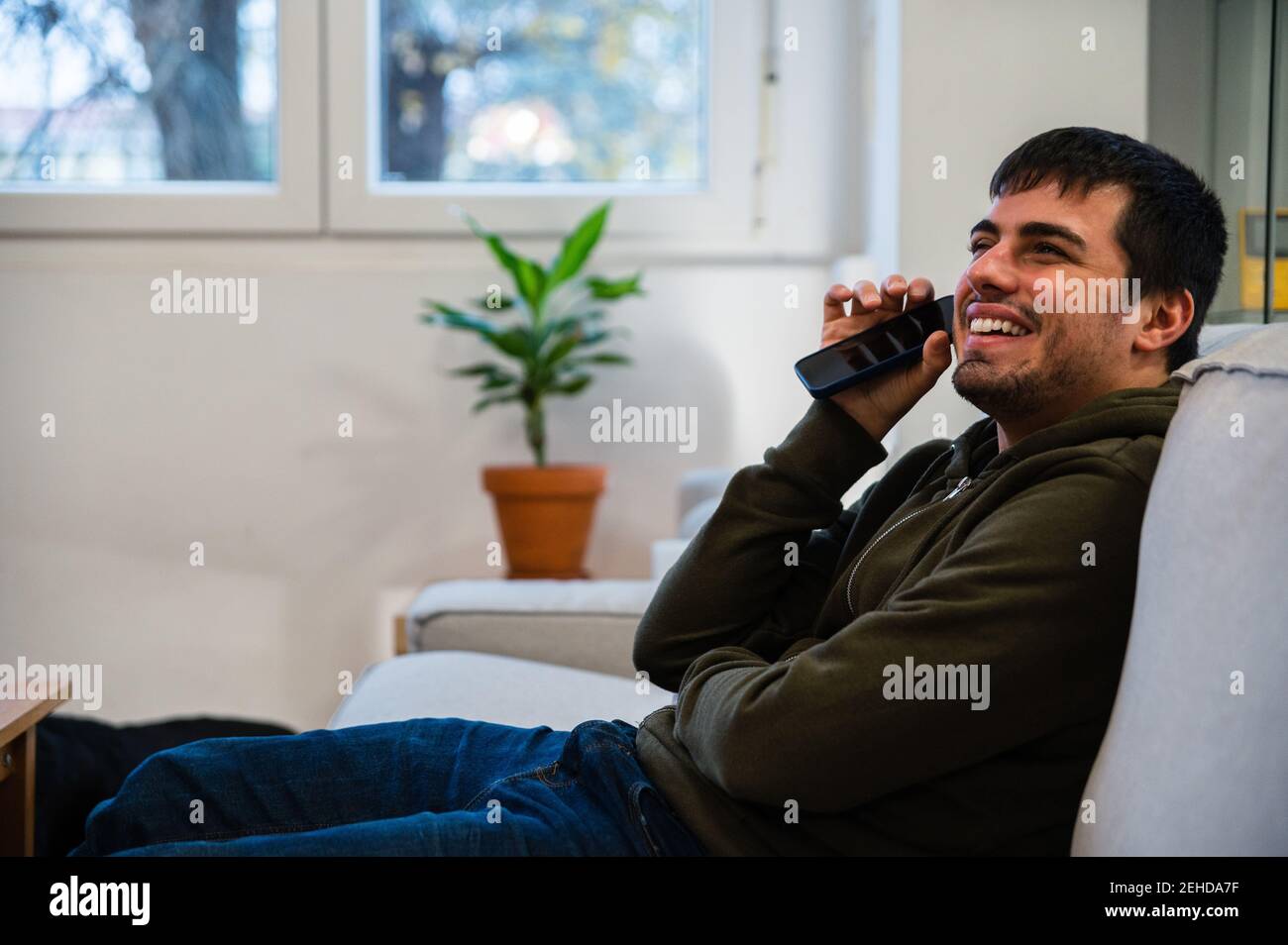 Vista laterale di un giovane maschio sorridente che soffre di cecità seduto sul divano e ascolto di messaggi audio sul telefono cellulare Foto Stock