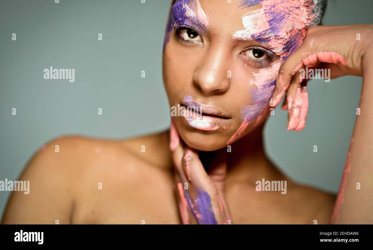 Modello etnico femminile creativo con viso cosparso di rosa e. vernice bianca appoggiata sulla mano e guardando la fotocamera sfondo grigio in studio Foto Stock