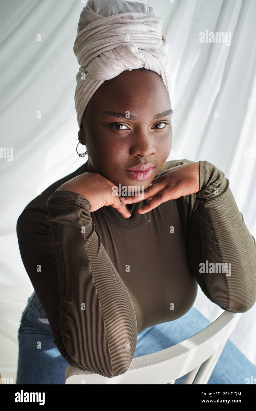 Dall'alto di seducente giovane donna nera in abiti casual e turbante seduto su sedia con le mani al mento e. guardando la fotocamera Foto Stock