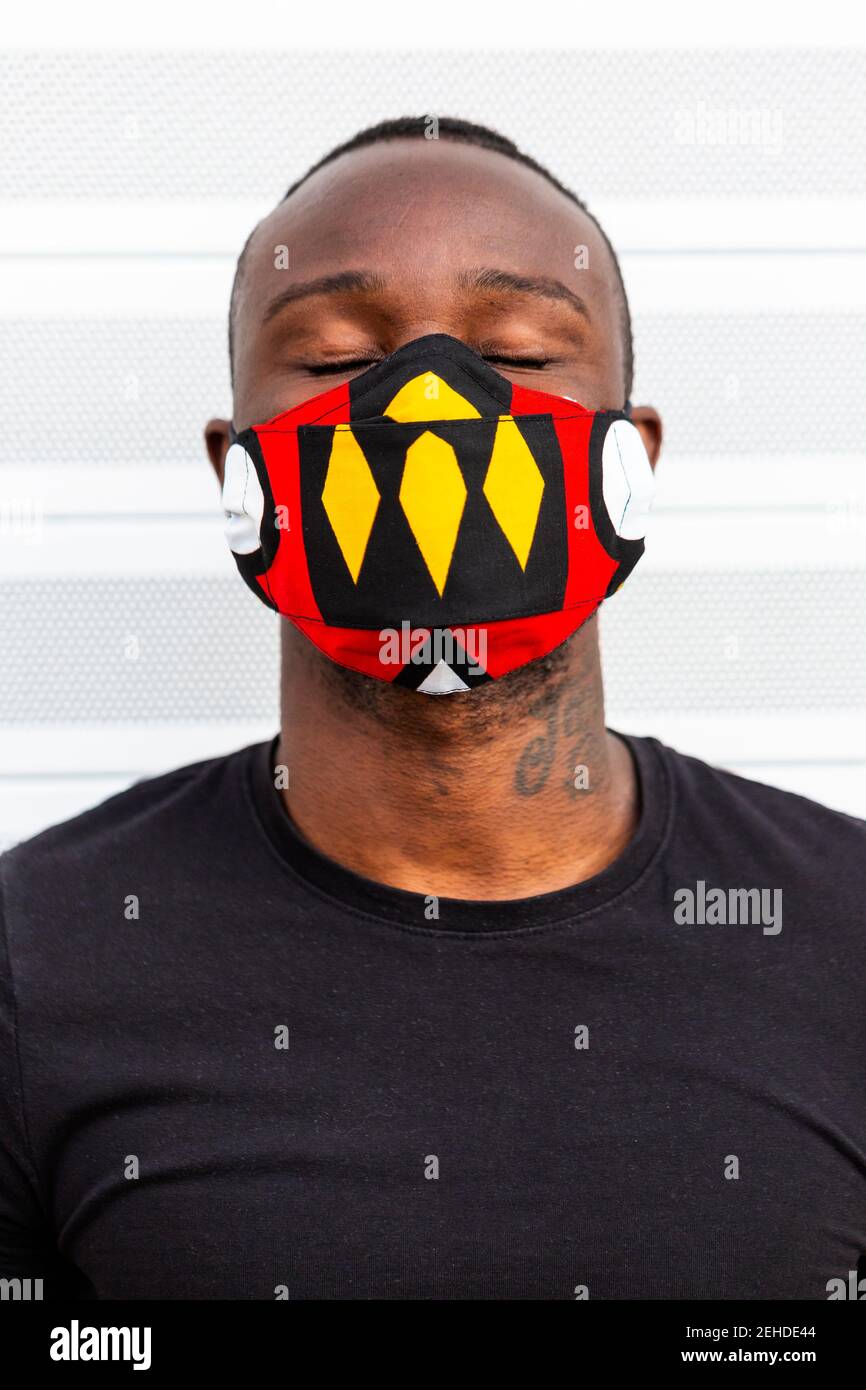 Anonimo giovane africano americano maschio con gli occhi chiusi in luminoso maschera con ornamento durante il periodo del coronavirus su sfondo chiaro Foto Stock