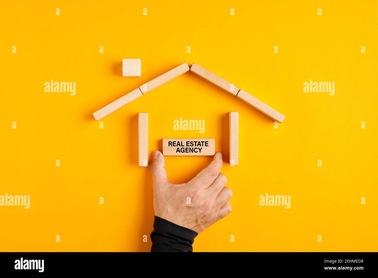 Mano maschio che mette il segno dell'agenzia immobiliare in una casa fatta di blocchi di legno su sfondo giallo. Settore immobiliare o di mercato, concetto di agenzia immobiliare. Foto Stock