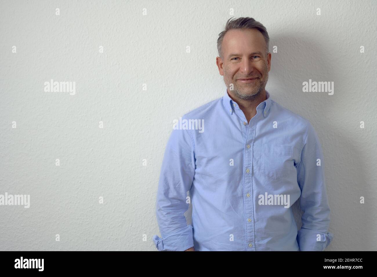 Bell'uomo maturo non rasato in camicia blu, in piedi contro la parete bianca e guardando la macchina fotografica con un sorriso amichevole. Ritratto frontale a mezza lunghezza con COP Foto Stock