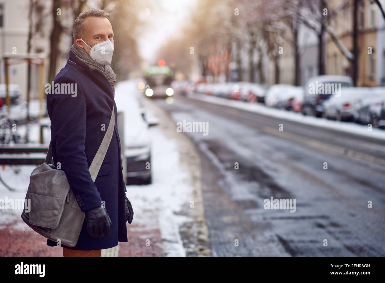 Donna bruna di mezza età in piedi a una fermata dell'autobus, indossando una maschera protettiva per il viso a causa del virus corona, in attesa del suo autobus per portarla al lavoro Foto Stock
