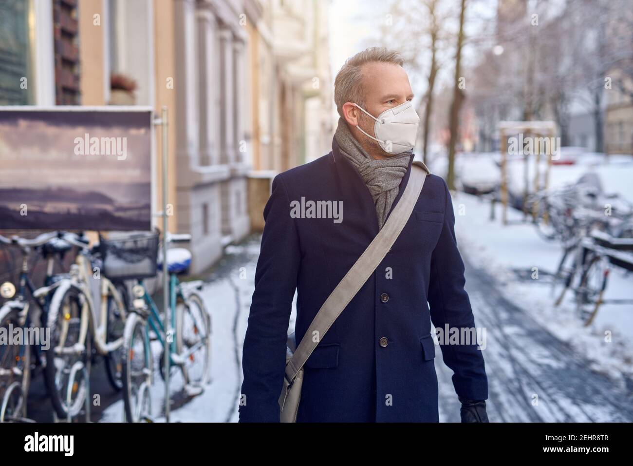 Uomo che indossa una maschera chirurgica protettiva durante il Covid-19 o coronavirus pandemic e overcoat invernale con borsa in pelle sopra la sua spalla che cammina Foto Stock