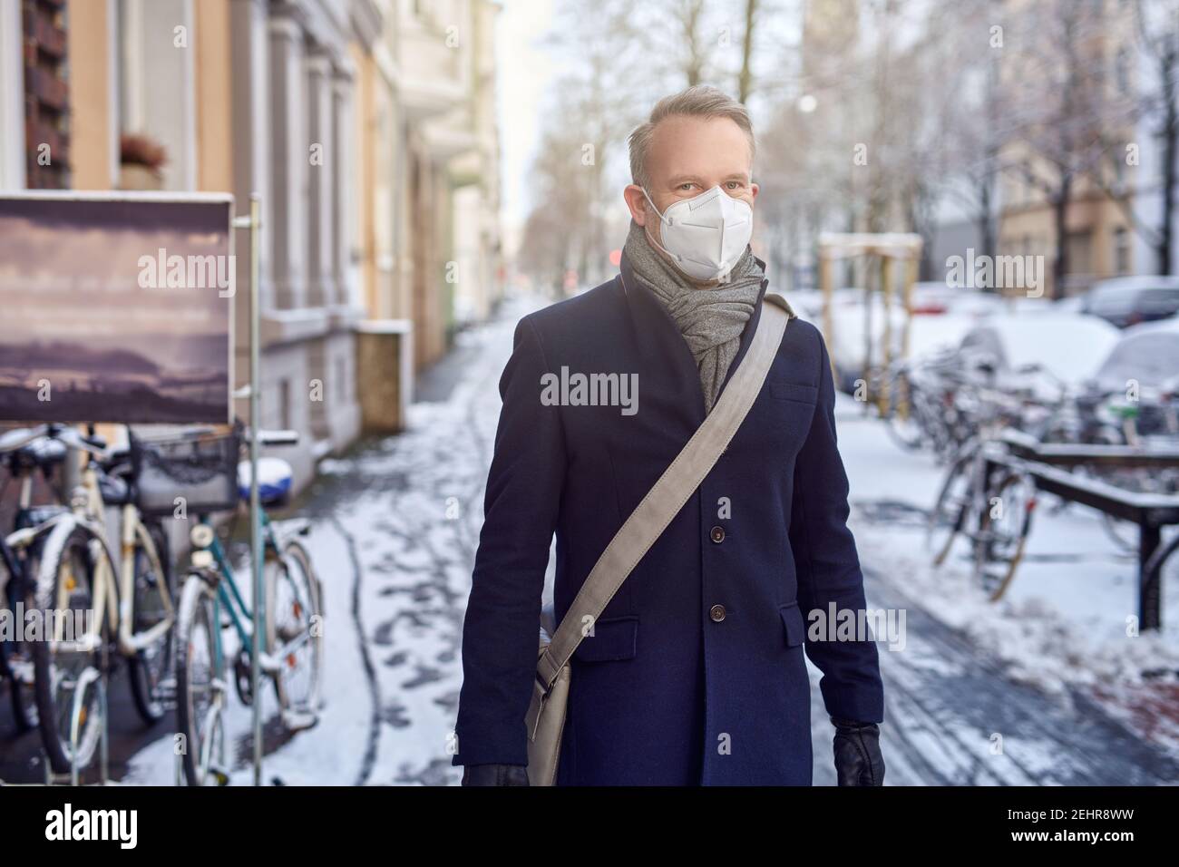 Uomo che indossa una maschera chirurgica protettiva durante il Covid-19 o coronavirus pandemic e overcoat invernale con borsa in pelle sopra la sua spalla che cammina Foto Stock