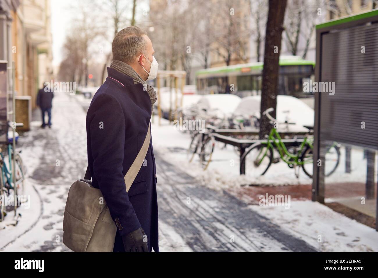 Uomo d'affari di mezza età in piedi a una fermata dell'autobus, indossando una maschera protettiva per il viso a causa del virus corona, in attesa del suo autobus per portarlo al lavoro Foto Stock