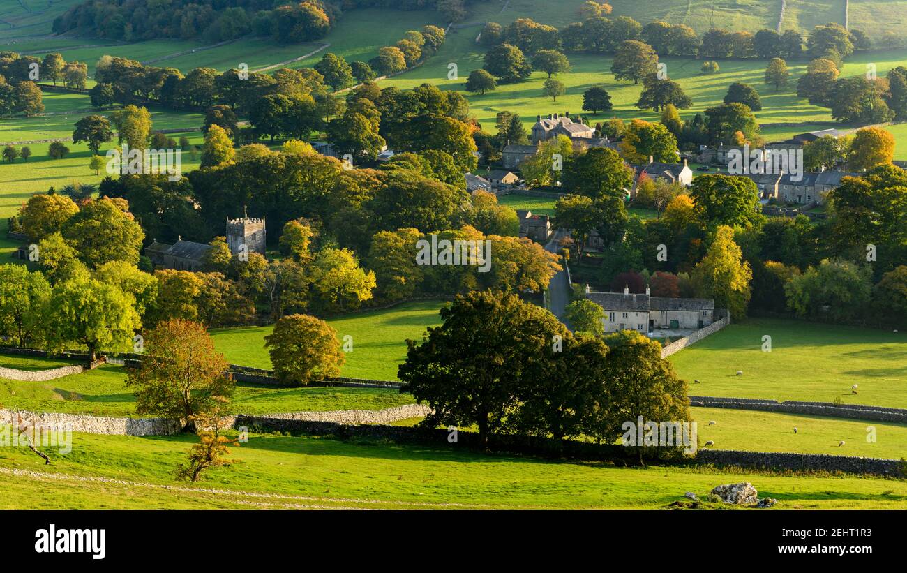 Sunlit pittoresco villaggio Dales (chiesa e case) annidato in valle da collina e alberi in colori autunnali - Arncliffe, Yorkshire Dales, Inghilterra, Regno Unito Foto Stock