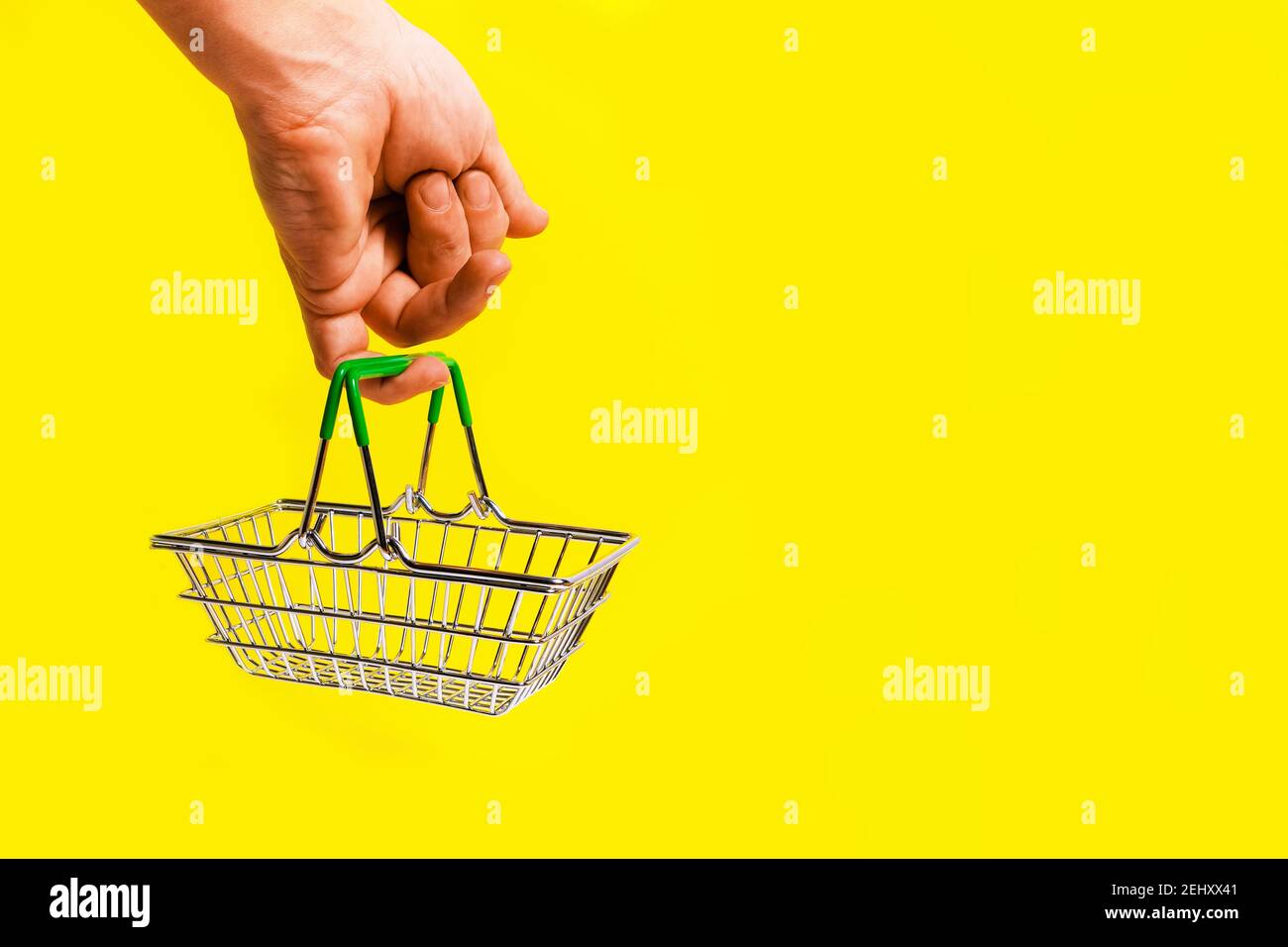 La mano tiene il cestino della spesa con il suo dito, su sfondo giallo. Concetto di un viaggio al supermercato. Foto Stock