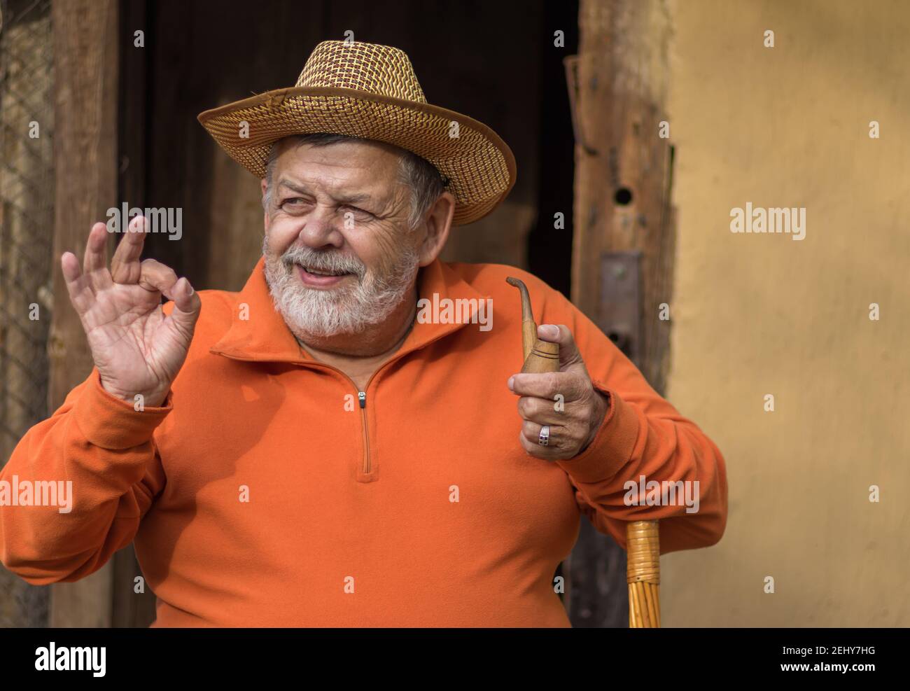 Ritratto di superiore caucasico positivo con tubo di tabacco che mostra il segno OK mentre si siede contro l'ingresso dell'antica casa Foto Stock