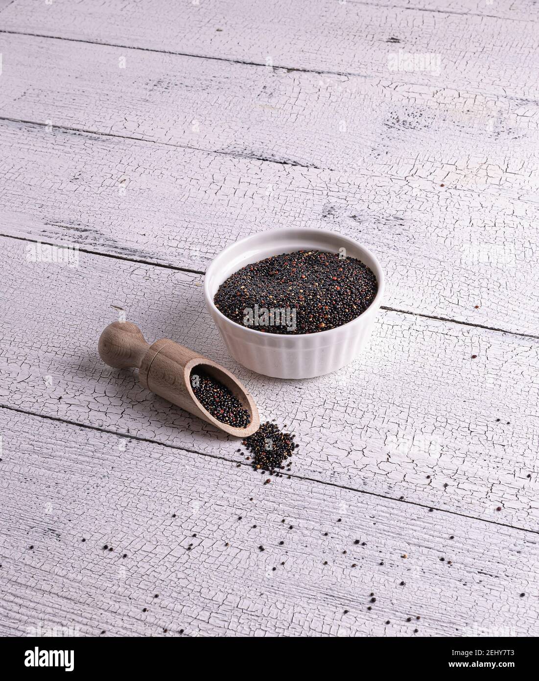 Semi crudi di quinoa neri non cotti in ciotola bianca e sccop con semi su tavolo di legno bianco. Pochi semi sono sparsi intorno. Cibo eccellente con spazio per la copia. Foto Stock