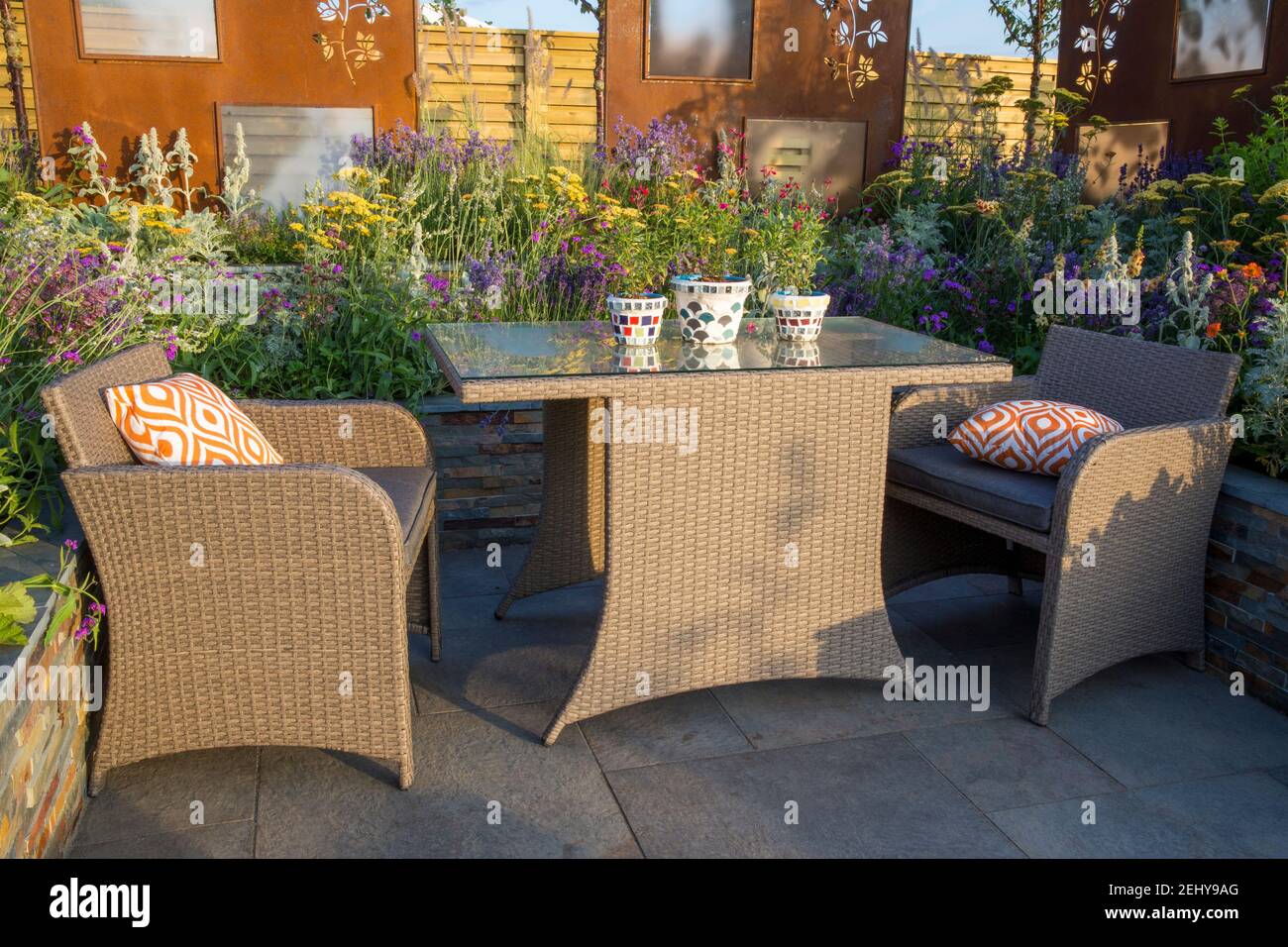 Patio dal design moderno con mobili da pranzo in rattan acrilico con tavolo e sedie per mobili da giardino, cuscini arancioni, letti rialzati UK Foto Stock