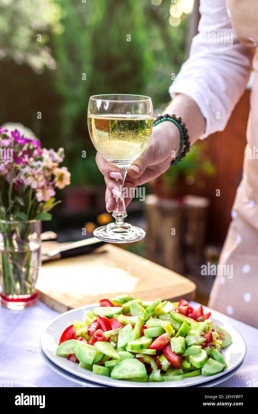 Donna che gusta il vino bianco durante la preparazione di insalata di verdure all'aperto. Preparazione per feste in giardino o celebrazioni Foto Stock