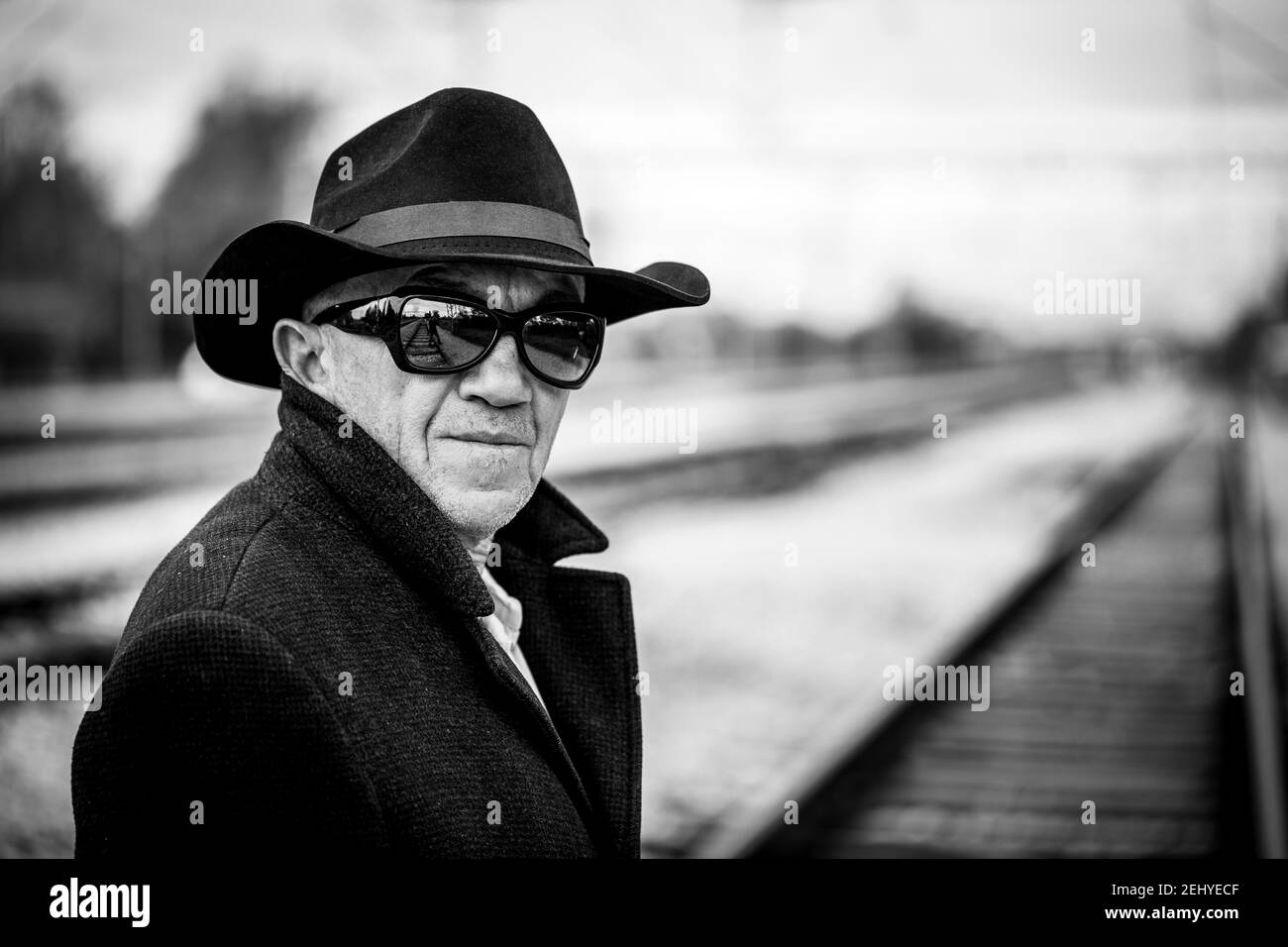 un uomo in un cappotto nero con un cappello nero e gli occhiali da sole neri  camminano lungo le piste ferroviarie Foto stock - Alamy