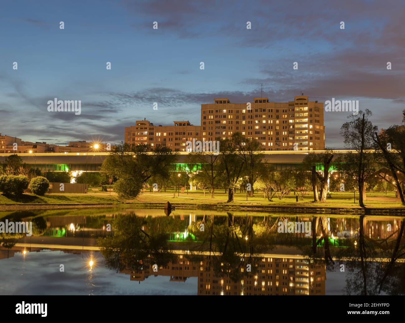 Paesaggio urbano serale. Case illuminate, sentieri nel parco della città, traccia sfocata del treno riflesso nel laghetto della città contro il cielo luminoso della sera Foto Stock
