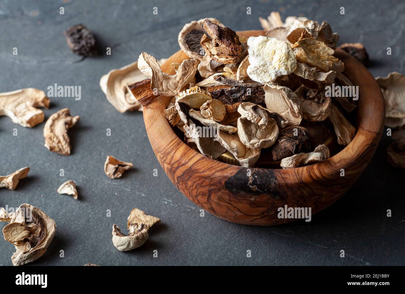 Primo piano immagine ad angolo isolata di una ciotola rustica di legno riempita con pezzi di funghi tagliati e secchi. Il recipiente si trova su una superficie di pietra scura. C'è un AS Foto Stock